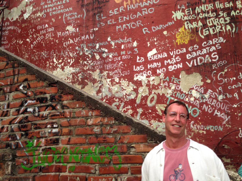 A smiling traveler stands before a red wall covered in graffiti