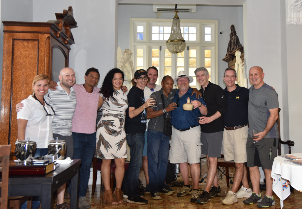 A dozen people pose for a group photo. The setting is indoors in a tall room with antique furniture and a window in the background. The man at the center of the group holds a ceramic jug of rum.