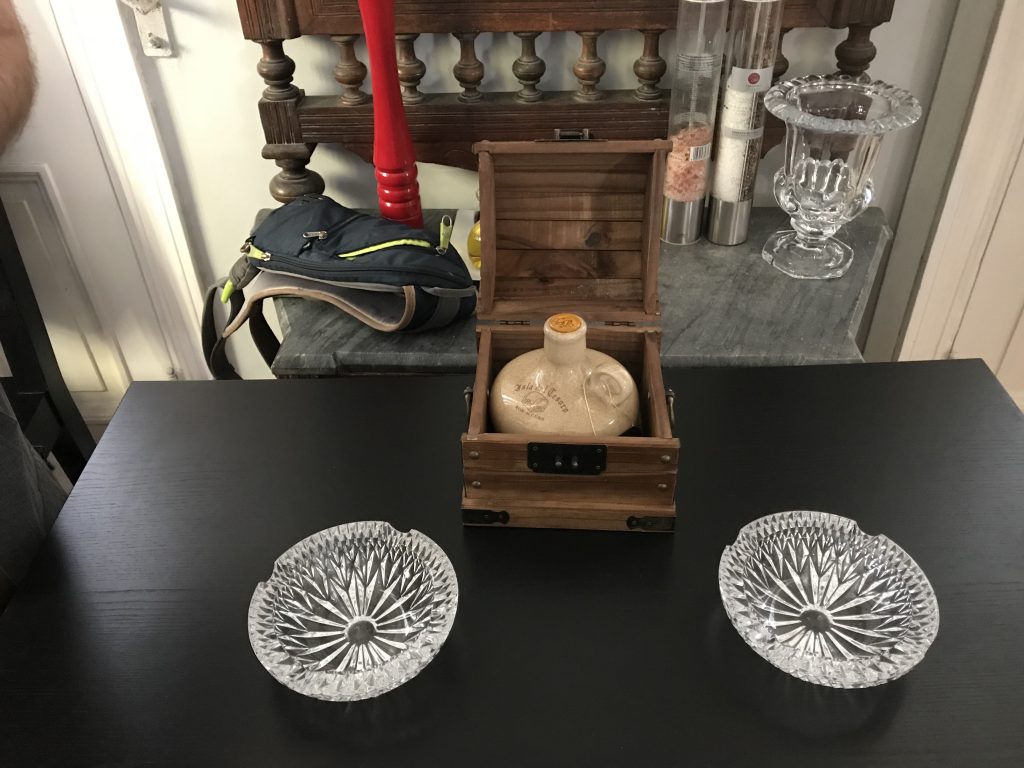 A small, open treasure chest contains a beige ceramic jug. On the table next to it are crystal ashtrays.