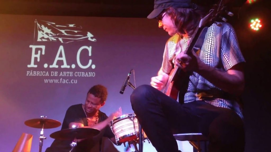 Foreground, a man sits on a stool playing an electric guitar. Background, a man sits at a drum set playing bongos. On the wall behind them is the logo for the Cuban Art Factory.