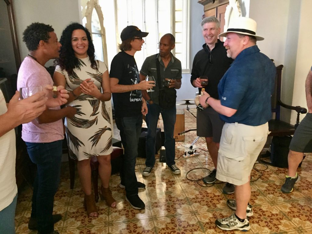 Six people stand chatting, five men and one woman, of varied races. The men are dressed fairly casually, and the woman is more dressed up in a white and gray print dress.