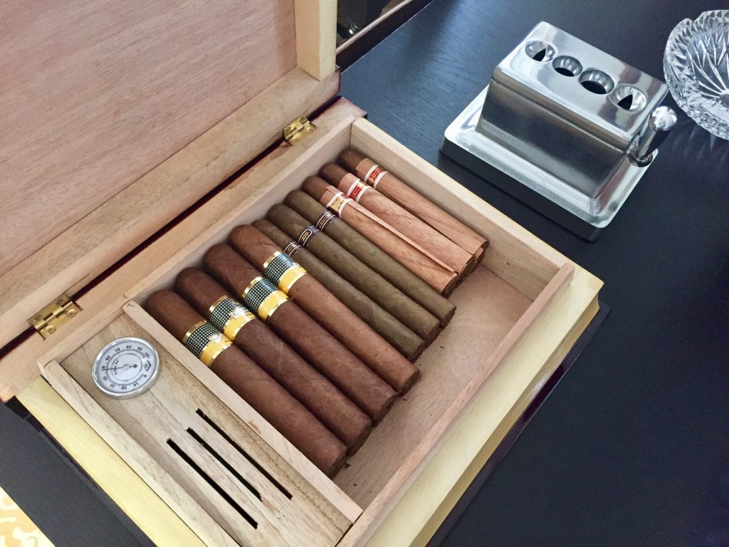 A humidor of cigars sits on a black tabletop alongside a stainless steel cigar cutter and crystal ashtray