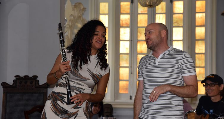 A woman and man stand side by side, angled toward one another, singing. The woman is dressed in a white and gray print dress and holds a clarinet in one hand. The man is wearing a white and gray striped polo. In the background is a window of yellow-orange stained glass.